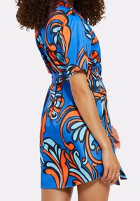 Helles blaues Kleid mit kräftigen orangen und türkisfarbenen wirbelnden Mustern, kurzen Ärmeln, einer taillierten Taille und einer weichen, glänzenden Stoffoberfläche.