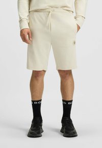 Lichtbeige shorts van zacht materiaal, met een trekkkoord in de taille, zijzakken en een subtiel hexagonaal logo op de linker bovenbeen. Zwarte sokken en sneakers.