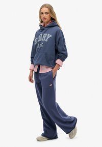 Mujer vistiendo una sudadera azul grande con el texto "GLORY N.Y.C", pantalones de chándal azules holgados, camiseta rosa debajo y zapatillas beige, de pie sobre un fondo blanco.