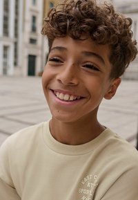 Light beige sweatshirt with ribbed neckline, featuring white text. Child with short, curly brown hair smiles against a blurred backdrop.