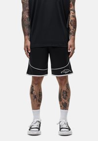 Zwarte sportshorts met witte piping, met een logo op het linkerbeen. Draagt witte crew-sokken en zwart-witte sneakers.