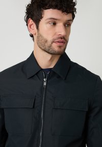 Hombre con cabello rizado y barba que lleva una chaqueta negra con cremallera y dos bolsillos en el pecho, mirando de lado contra un fondo claro y liso.