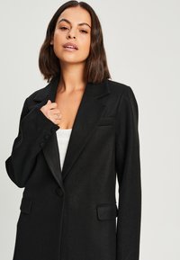 Blazer noir ajusté avec un col cranté, fermeture à un bouton et deux poches à l'avant. Texture douce et design structuré.
