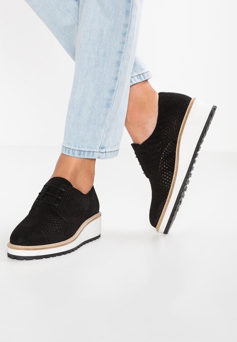 Scarpe oxford nere con texture, suola piatta bianca e grip in gomma nera, abbinate a jeans azzurri arrotolati.