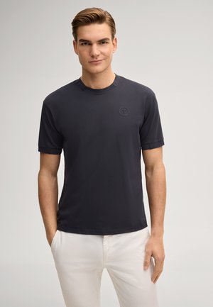 Junger Mann mit den Händen in den Taschen, trägt ein marineblaues kurzärmeliges Hemd und weiße Hose, steht vor einem schlichten hellen Hintergrund.