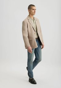 Blazer beige claro con solapas de muesca, bolsillos delanteros y botones dorados, combinado con una camisa verde estampada y jeans azules. Zapatos negros.