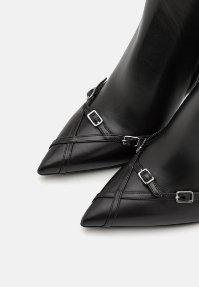 Botines de cuero negro con punta afilada, adornados con hebillas plateadas y un diseño de correas cruzadas, con una textura suave y una silueta elegante.