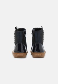 Bottes de cheville en cuir verni noir avec un dos texturé, des fermetures éclair sur les côtés et des semelles en caoutchouc marron. Présente des accents décoratifs sur les côtés.