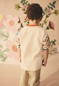 Vaikas dėvi baltą marškinėlį su spalvingomis raštuotomis rankovėmis ir oranžine apykakle, derančią su šviesiai rudos spalvos dryžuotomis kelnėmis. Fone matyti gėlių raštai.