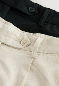 Zbliżenie na paski i guziki w beżowych i czarnych lnianych spodniach, pokazujące szlufki na pasek i przeszyte brzegi.