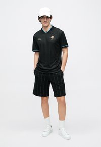 Νέος άνδρας που φοράει μαύρο αθλητικό πουκάμισο και σορτς EA7, λευκό καπέλο, λευκές κάλτσες και λευκά αθλητικά παπούτσια, στέκεται με τα χέρια στις τσέπες.