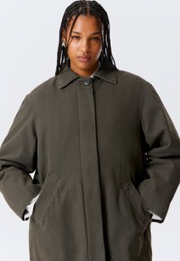 Grüner Baumwollmantel mit geradem Schnitt, großem Knopf, spitzem Kragen und Seitentaschen. Verfügt über eine strukturierte Oberfläche und eine lockere Passform.