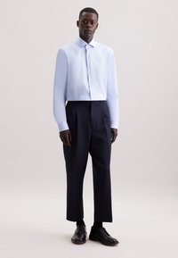 Camicia azzurra a maniche lunghe con colletto abbottonato, abbinata a pantaloni blu navy plissettati e scarpe nere con lacci. Texture morbide in tutto.