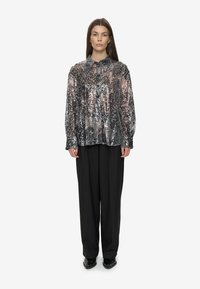 Femme portant une blouse en sequins argentés à boutons et manches longues, associée à un pantalon noir ample et des chaussures à talons noirs, se tenant droite.