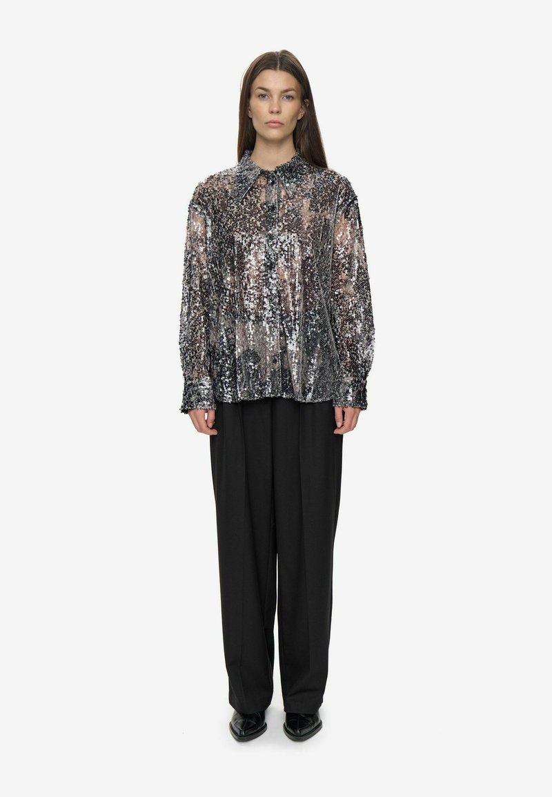 Femme portant une blouse en sequins argentés à boutons et manches longues, associée à un pantalon noir ample et des chaussures à talons noirs, se tenant droite.