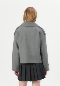 Giacca di lana grigia con colletto a punta, vestibilità ampia e spalline, abbinata a una gonna plissettata scura. Design semplice e strutturato.