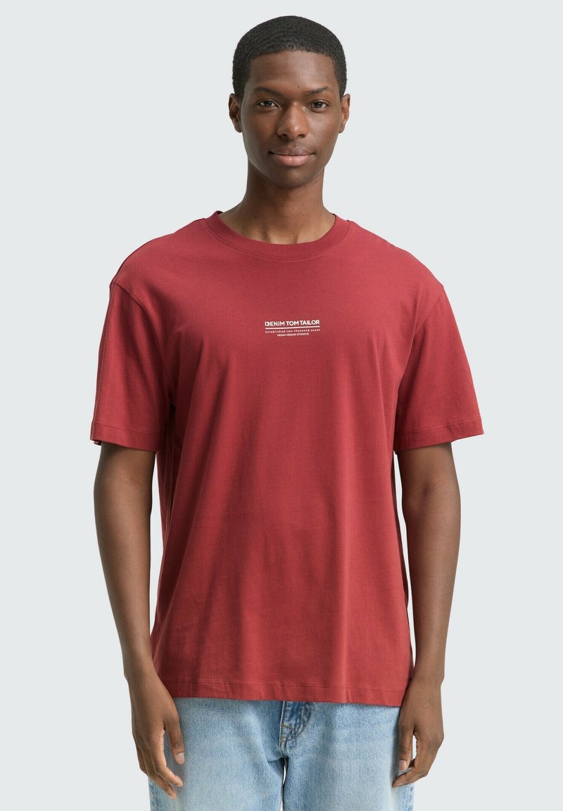 Mężczyzna w gładkiej czerwonej koszulce z małym białym napisem na klatce piersiowej i jasnoniebieskich dżinsach, stojący na jednolitym jasnym tle.