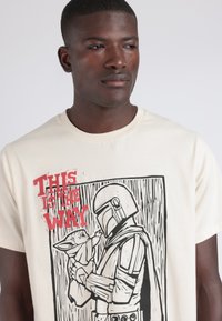 Camiseta color crema con una impresión gráfica de un personaje con un casco sosteniendo una figura pequeña, con el texto rojo "ESTE ES EL CAMINO."