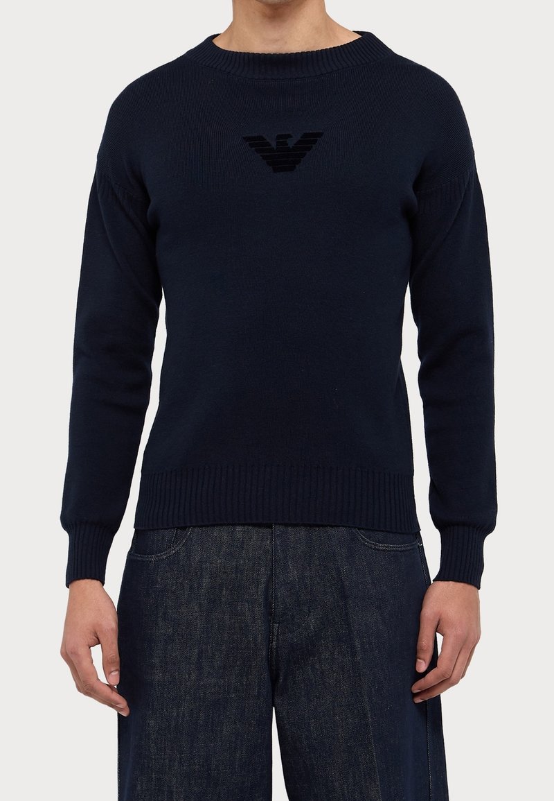 Homme portant un pull bleu marine à manches longues avec un emblème noir sur la poitrine et un pantalon en jean bleu foncé, devant un fond uni.
