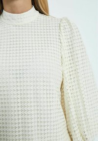 Spetsblus i off-white med hög hals, strukturerat mönster och puffärmar, som visar intrikata geometriska mönster och mjukt tyg.