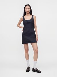 Femme aux longs cheveux noirs portant une mini-robe noire sans manches, des chaussettes blanches et des mocassins noirs, debout avec une main sur la hanche.