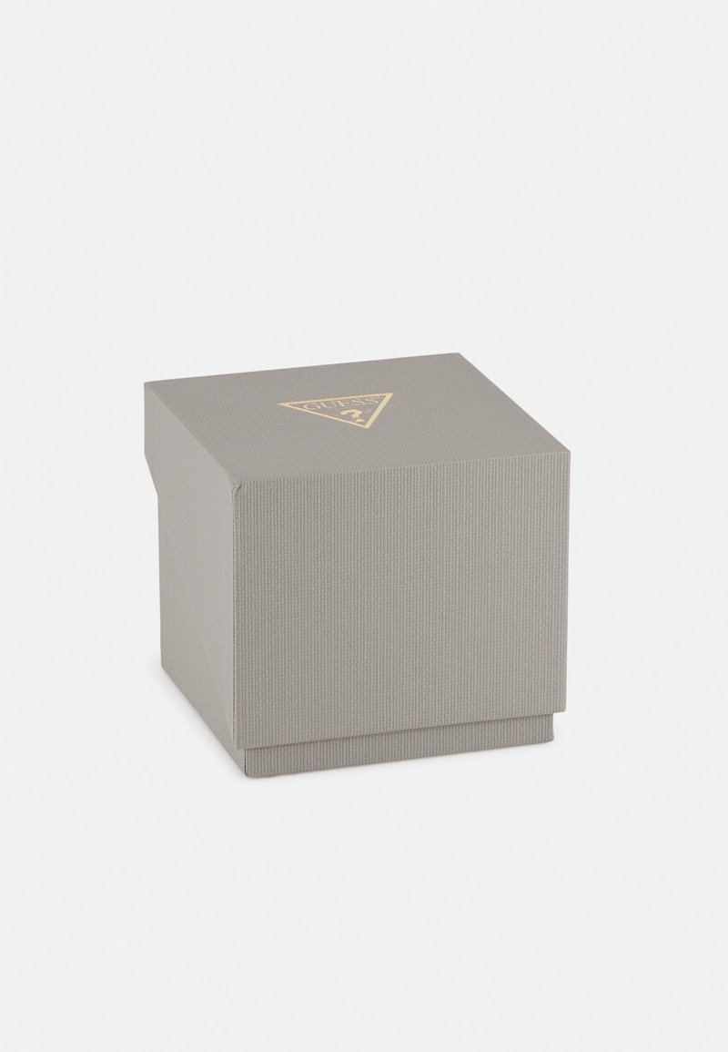 Boîte carrée texturée grise avec un logo triangulaire doré Guess sur le dessus, placée sur un fond blanc uni.