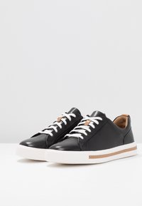 Baskets en cuir noir avec lacets blancs, semelle en caoutchouc blanche, accents tan et un design minimaliste. Texture lisse avec une forme arrondie.