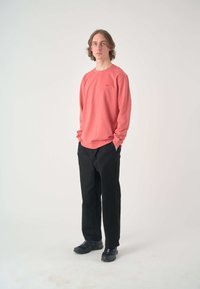 Long-sleeve, coral sweatshirt with a crew neck, featuring a small logo, paired with black, loose-fit trousers and black athletic shoes.