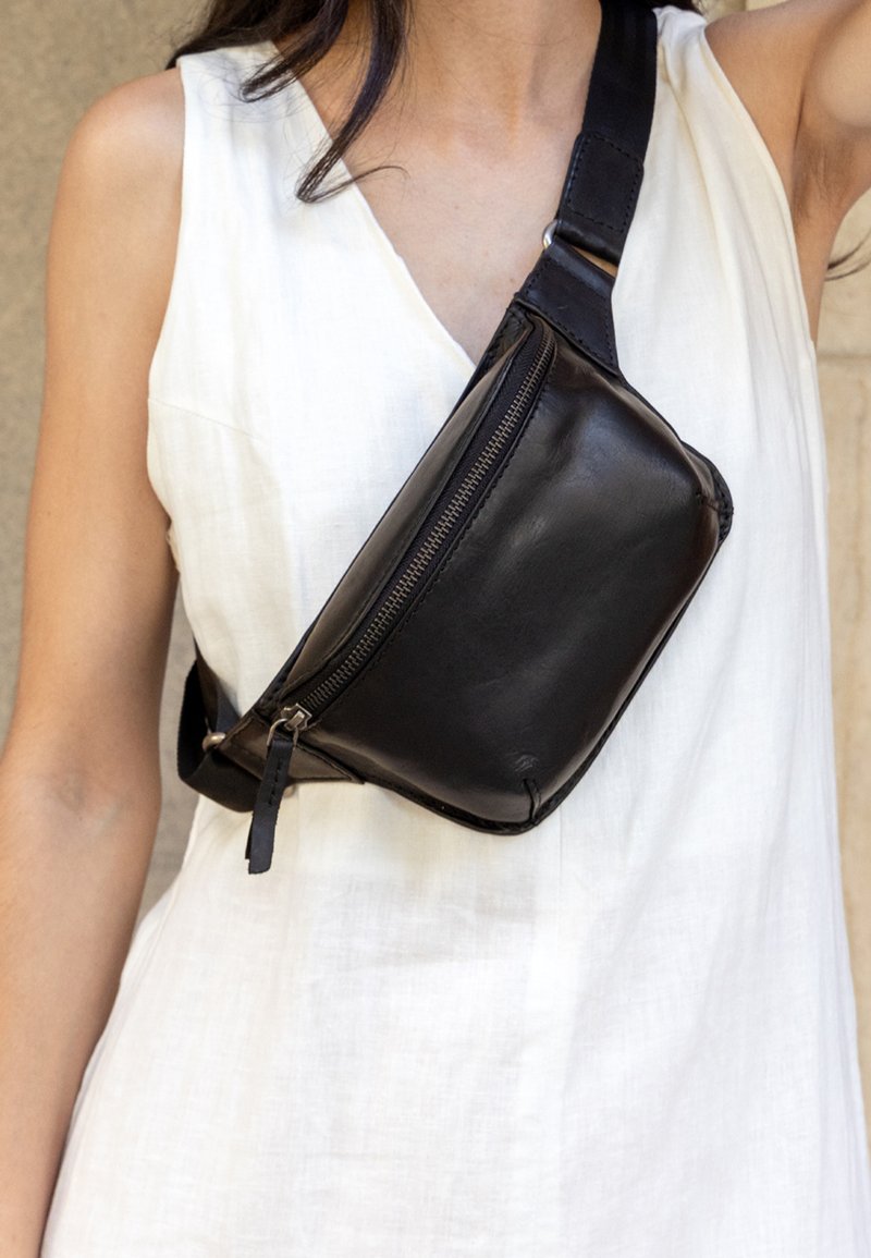 Borsa a tracolla nera in pelle sintetica con una leggera lucentezza, dotata di un compartimento con zip e una tracolla regolabile, indossata sopra un capo bianco.