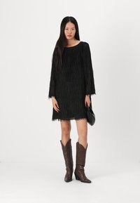 Abito nero con frange, maniche lunghe, scollatura rotonda e rifinitura testurizzata, abbinato a stivali marroni al ginocchio e una borsa nera.