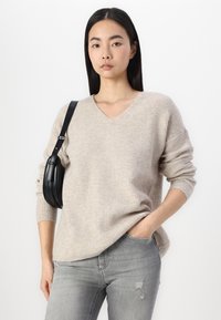 Beige geripptes Strickpullover mit V-Ausschnitt, langen Ärmeln und lockerer Passform, kombiniert mit hellgrauen, distressed Jeans und einer schwarzen Umhängetasche.