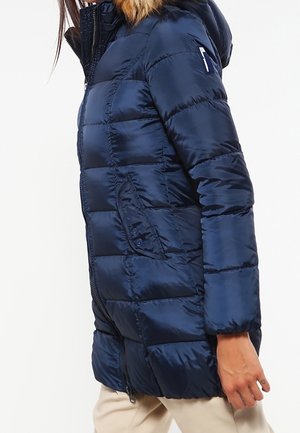 Kobieta w granatowej pikowanej kurtce puchowej z kapturem i przednimi kieszeniami z patkami, w połączeniu z beżowymi spodniami.