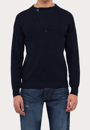 Hombre con suéter de punto azul marino oscuro con solapa abotonada en el cuello y vaqueros azules con ligero desgaste en el muslo.