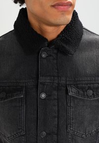 Svart jeansjacka med en fluffy svart krage, med framfickor, knäppning med knappar och en texturerad tygdesign.