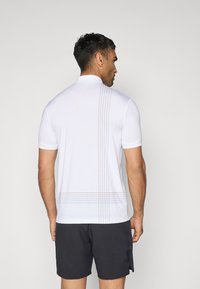 Kurzärmliges Poloshirt in Weiß, mit strukturiertem Oberflächendesign und vertikalen Linienakzenten in Grau und Orange auf dem Rücken. Dazu schwarze Shorts.