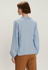 Jasnoniebieska bluzka z długimi, bufiastymi rękawami i kołnierzykiem z tyłu. Posiada pionowe paski i luźny krój. Zestawiona z brązowymi spodniami.