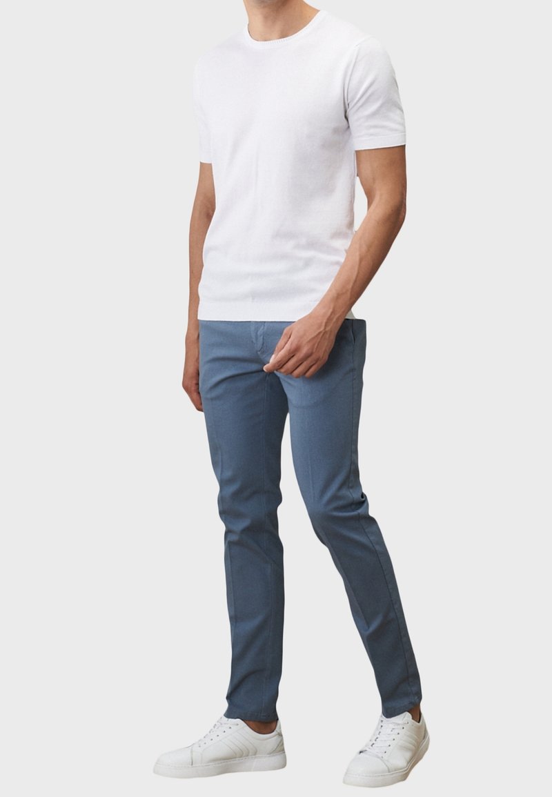 Homme portant un t-shirt blanc à manches courtes, un pantalon slim bleu et des baskets blanches, debout devant un fond uni.
