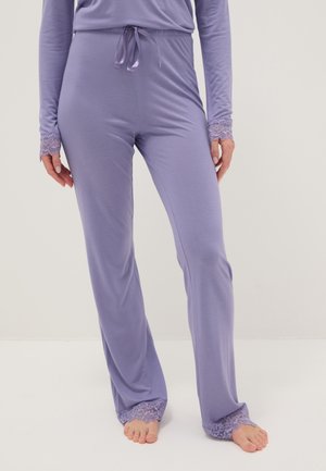 Donna che indossa una maglia lavanda a maniche lunghe e pantaloni abbinati con bordi in pizzo sui polsini e sulle gambe dei pantaloni, in piedi a piedi nudi su sfondo semplice.