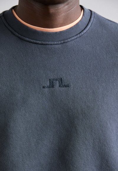 Sweatshirt cinza com gola canelada, apresentando um logótipo em relevo discreto no peito. O material parece suave e texturizado.