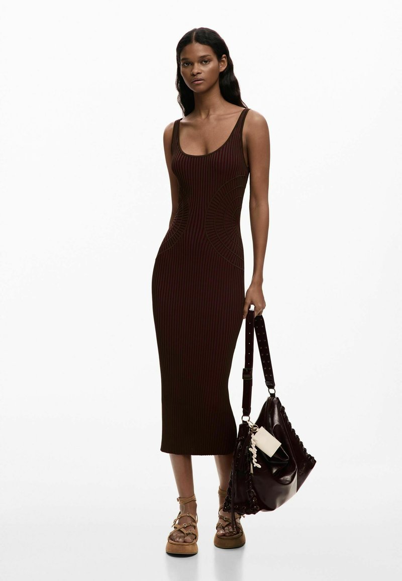 Kobieta ubrana w dopasowaną, ciemnobrązową, prążkowaną sukienkę midi bez rękawów, trzymająca dużą ciemną torebkę i mająca na sobie cieliste sandały na platformie z paskami.