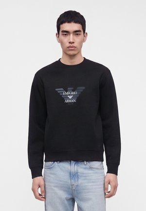 Młody mężczyzna w czarnej bluzie Emporio Armani z logo i jasnoniebieskich dżinsach, stojący na tle jednolitego jasnego tła.
