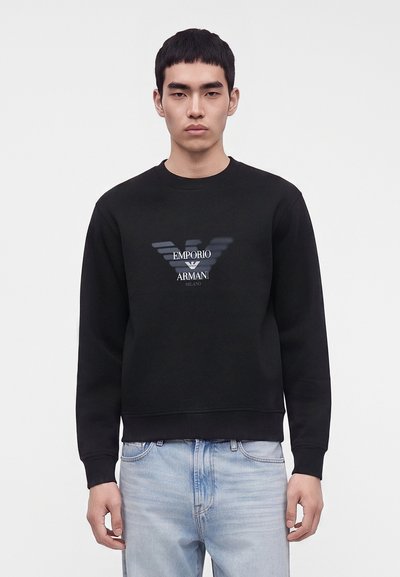 Млад мъж, носещ черен суичър Emporio Armani с лого и светлосини дънки, стои на фона на едноцветен светъл фон.