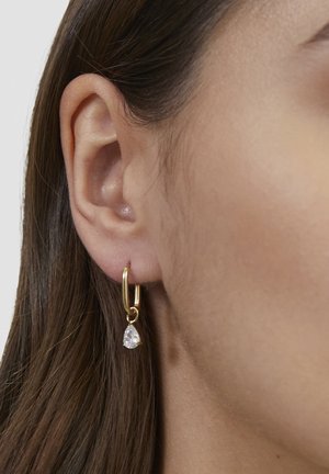 Gold hoop earring with a pear-shaped clear gemstone pendant, displayed against skin, highlighting the earring's design and texture.