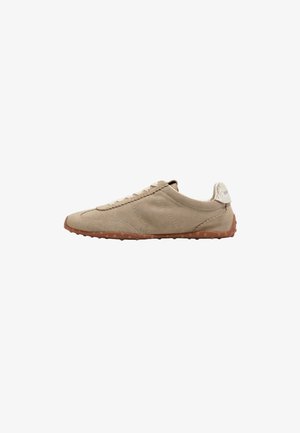 Beige suède sneaker met een gestructureerd oppervlak, vetersluiting en een bruine rubberen zool met kleine stippen voor grip. De achterkant heeft een lichtgekleurde accent.