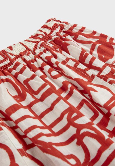 Bijela tkanina s širokim crvenim apstraktnim uzorcima linija, s elastičnim pojasom i vidljivim šavovima.