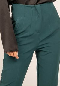 Pantalons taille haute teal avec poches avant, détails plissés et texture de tissu lisse, associés à un haut noir à manches longues.