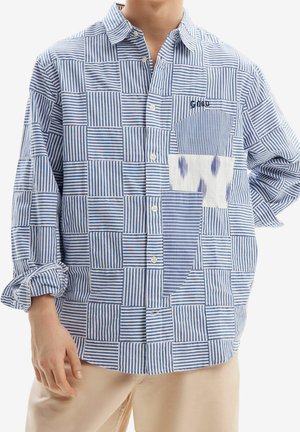 Homme portant une chemise boutonnée rayée bleu et blanc en patchwork avec manches retroussées et pantalon beige, debout devant un fond blanc.