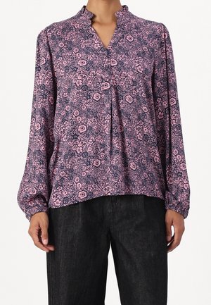 Langærmet lyserød bluse med blomsterprint og flæsekant på kraven, båret med sorte bukser på en person vist fra nakke til hofter.