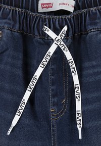 Mörkblå denimjeans med elastisk midja, med ett vitt snöre med "LEVI'S"-logotyp och gula söms detaljer.
