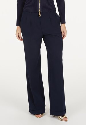 Frau trägt marineblaue, weit geschnittene Hosen mit vorderen Falten und Taschen, kombiniert mit einem marineblauen, gerippten Zip-Up-Top und goldenen offenen High Heels.